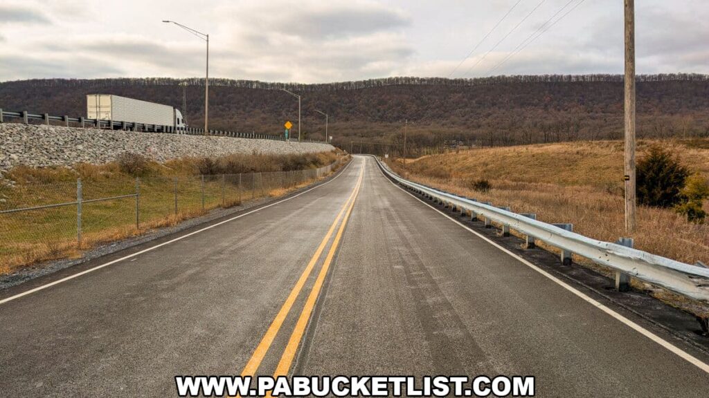 Two-lane road leading from the Sideling Hill Service Plaza toward the trailhead for the 8.5-mile Abandoned PA Turnpike, with guardrails, open fields, and a ridgeline rising in the distance along the Bedford–Fulton county line.