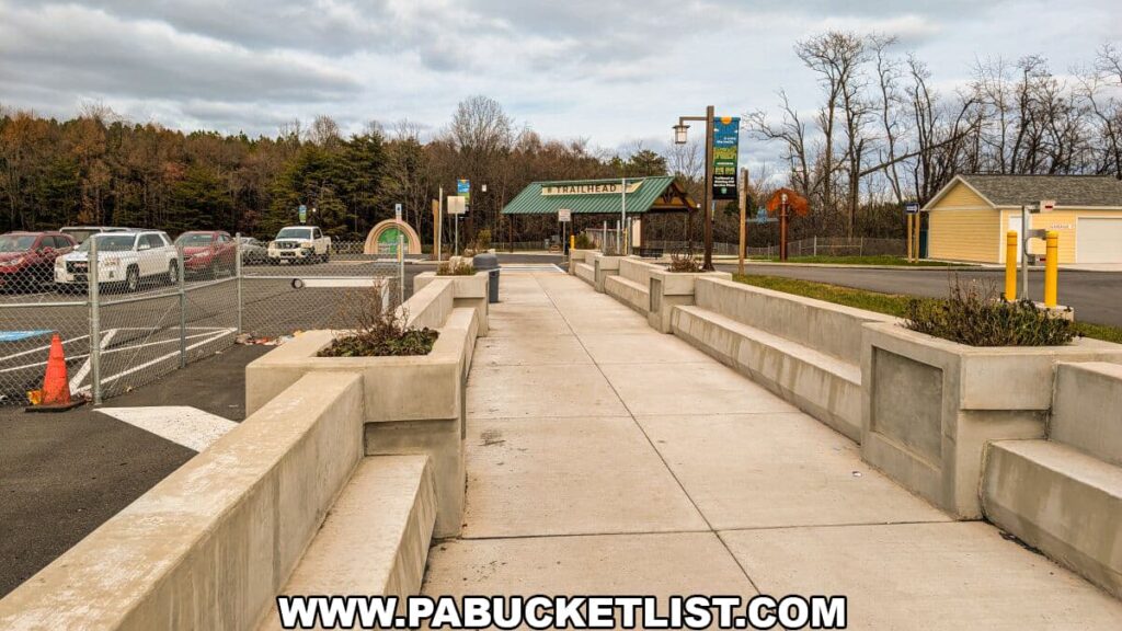 Concrete walkway leading from the Sideling Hill Service Plaza toward the covered trailhead pavilion, with parking areas, signage, and surrounding woodland marking the access point to the 8.5-mile Abandoned PA Turnpike in Bedford and Fulton counties.