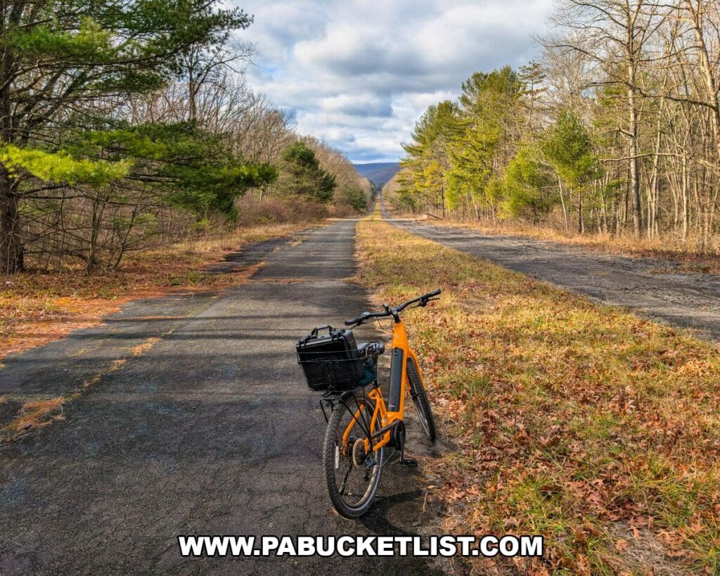 An orange bicycle sits on the cracked, weathered pavement of the Abandoned PA Turnpike, with the long, straight roadway disappearing into the distance between evergreen stands and leafless hardwoods along this 8.5-mile former Turnpike corridor in Bedford and Fulton counties.