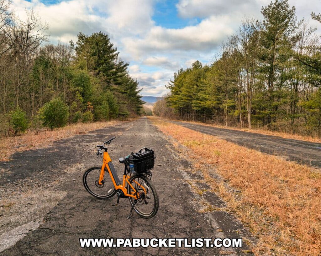 An orange bicycle stands on the cracked, overgrown pavement of the Abandoned PA Turnpike, with the long, tree-lined roadway stretching toward distant ridges under a partly cloudy sky along this 8.5-mile former Turnpike route in Bedford and Fulton counties.