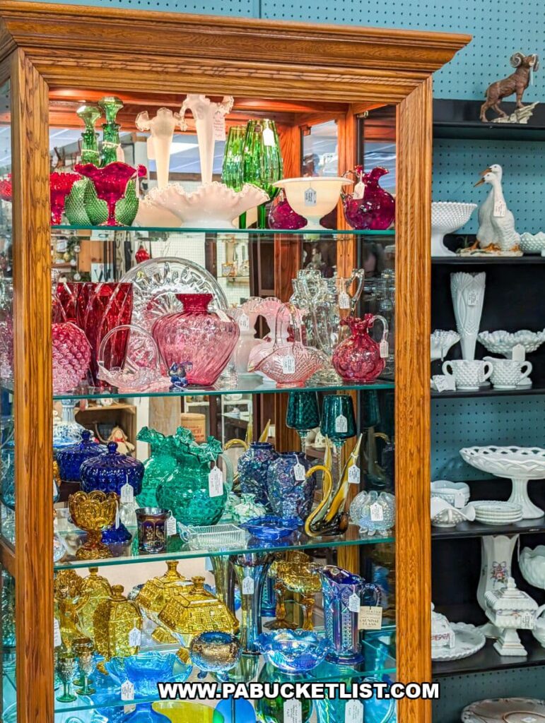 Glass display cabinet at Black Rose Antiques in Hanover, Pennsylvania, filled with colorful vintage glassware including vases, pitchers, bowls, and decorative pieces in shades of pink, green, blue, and gold, with additional white ceramic items on nearby shelves.