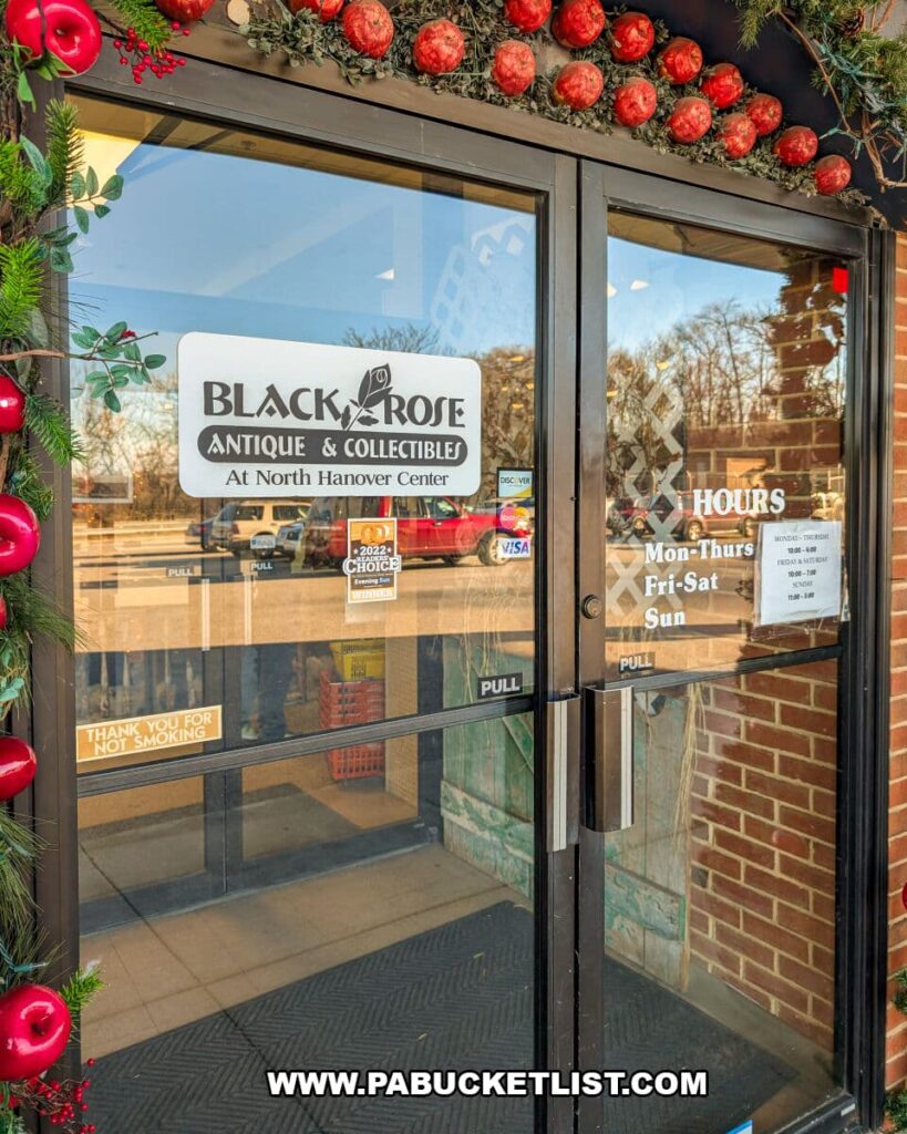 Front entrance doors of Black Rose Antique & Collectibles at North Hanover Center in Hanover, Pennsylvania, decorated with a garland of red ornaments and greenery, with business hours and signage visible on the glass.