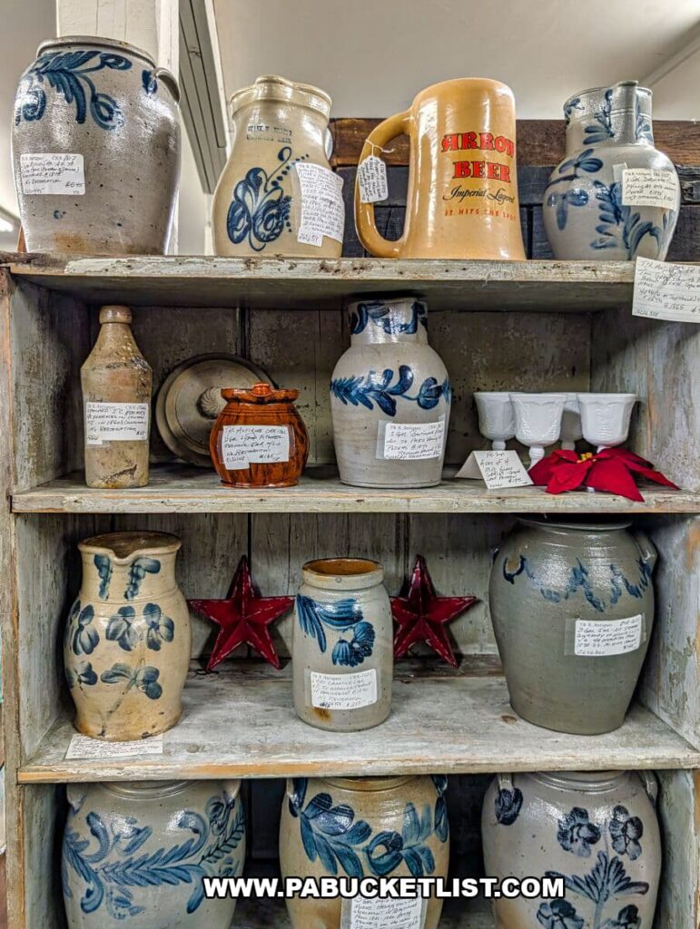 Display of antique stoneware crocks and jugs with cobalt blue floral designs arranged on rustic wooden shelves at Collector’s Choice Antiques Gallery in New Oxford, Pennsylvania, alongside vintage pottery pieces, red star décor accents, and an old Arrow Beer stein.