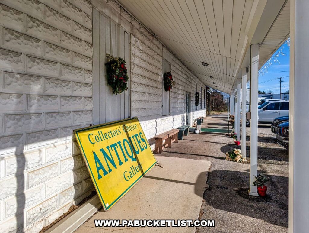 Side exterior walkway of Collector’s Choice Antiques Gallery in New Oxford, Pennsylvania, featuring a large yellow antiques sign propped against the white brick building, holiday wreath décor, and parked cars nearby.