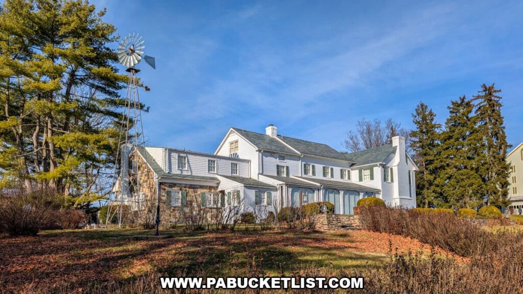 Exterior view of the white-painted farmhouse at the Eisenhower National Historic Site in Gettysburg, surrounded by winter-bare landscaping and tall evergreens, with a metal windmill rising beside the stone and wood structure under a bright blue sky.