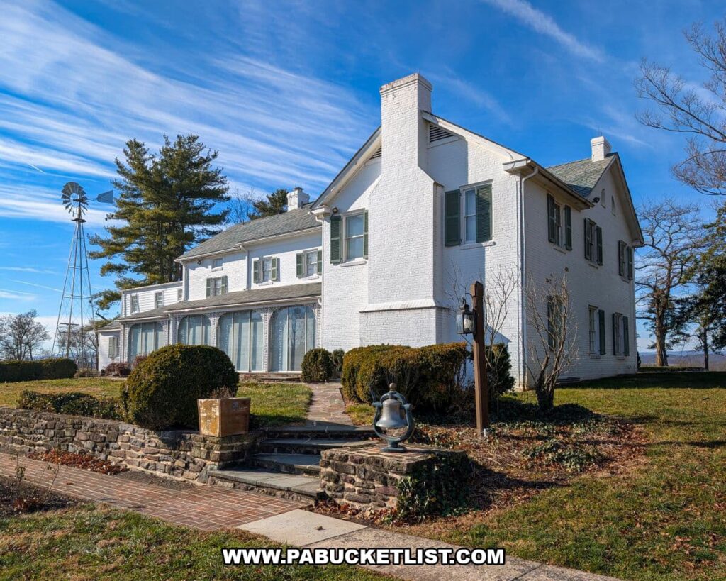 Back view of the white-painted farmhouse at the Eisenhower National Historic Site, showing its long porch, stone walkway, manicured shrubs, a metal windmill, and expansive blue-sky landscape in Gettysburg.