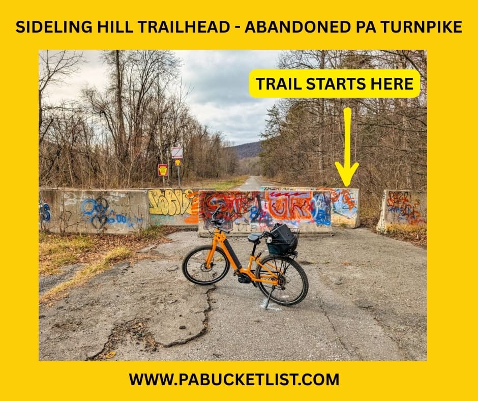 Photo of the Sideling Hill Trailhead entrance to the Abandoned PA Turnpike, featuring an orange bicycle in front of graffiti-covered concrete barriers and a marked pathway showing where the 8.5-mile former Turnpike trail begins in Bedford and Fulton counties.
