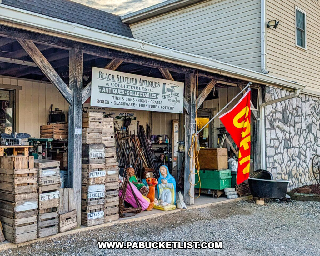 The covered entrance of Black Shutter Antiques in New Oxford, Pennsylvania, featuring stacked wooden crates, vintage tools, religious statuary, an OPEN flag, and architectural salvage displayed beneath a rustic porch at the multi-vendor antique store