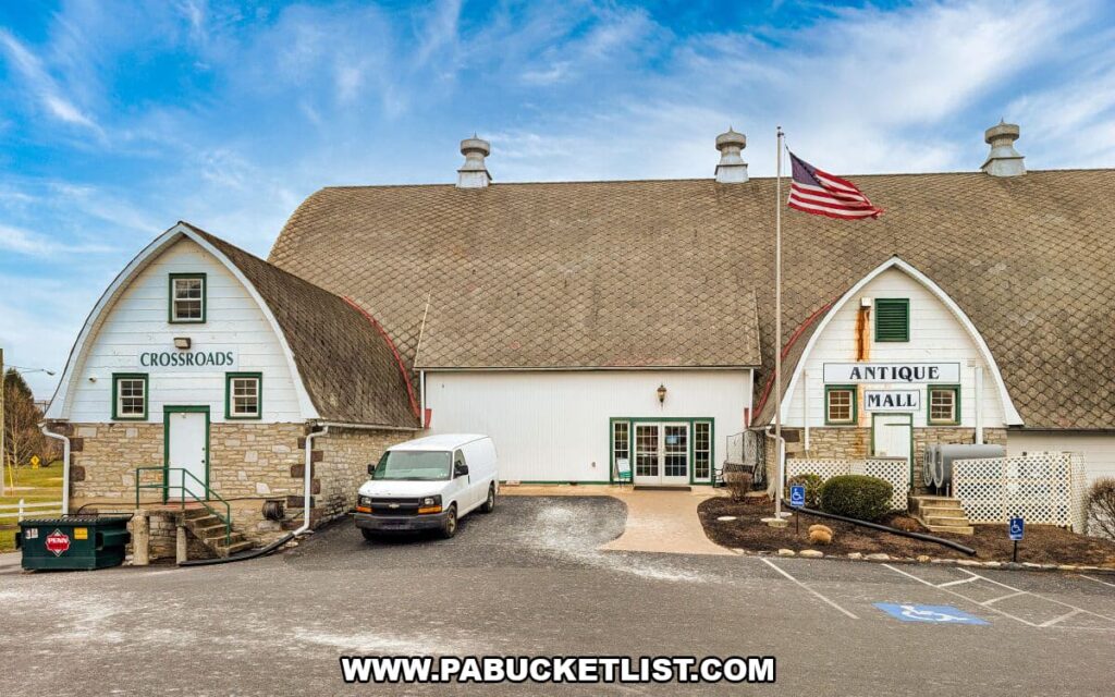 Exterior view of Crossroads Antique Mall in Hershey, Pennsylvania, showing the large historic barn with gambrel rooflines, white siding and stone accents, antique mall signage, and an American flag flying above the entrance.