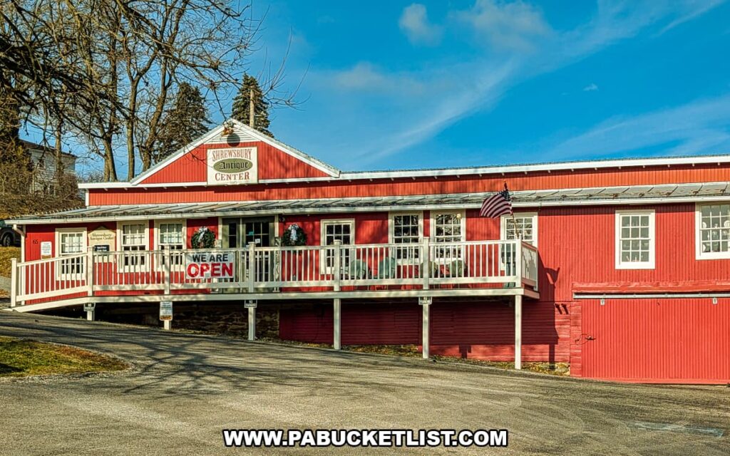 Exterior view of the red barn-style Shrewsbury Antique Center building with a covered porch, storefront sign, and American flag on a sunny day in York County, Pennsylvania.