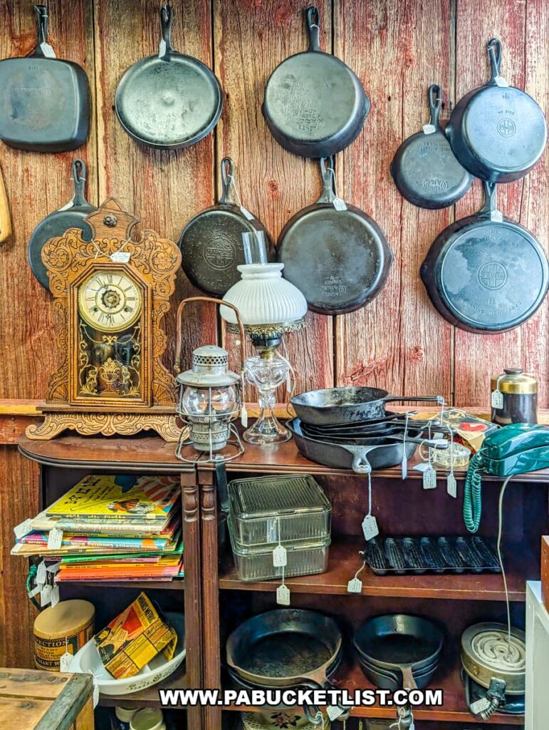 Display of vintage cast iron skillets hanging on a wooden wall above shelves filled with antique cookware, lanterns, clocks, and kitchen collectibles inside Shrewsbury Antique Center in York County, Pennsylvania.