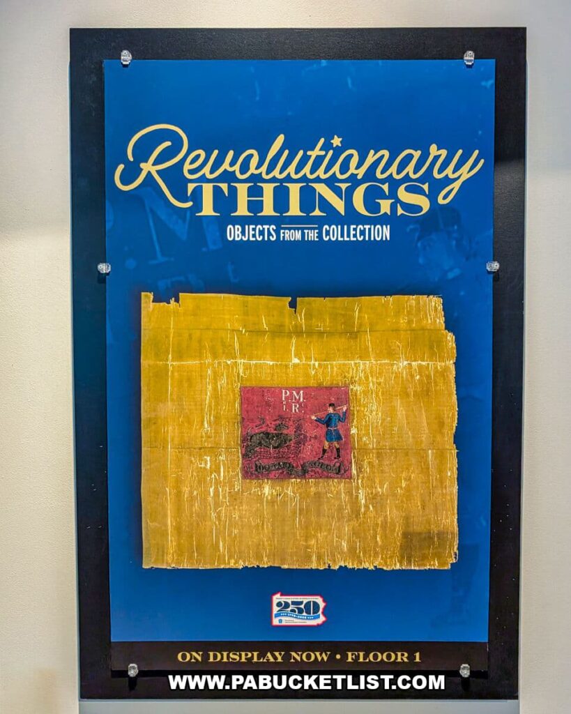 Poster at The State Museum of Pennsylvania in Harrisburg featuring the Revolutionary Things exhibit, with a blue background, gold lettering, and a historic Revolutionary War–era object mounted at the center, highlighting artifacts from Pennsylvania’s colonial past on display on the museum’s first floor