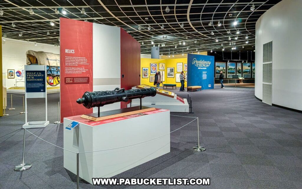 Wide view of the Revolutionary Things exhibit on the first floor of The State Museum of Pennsylvania in Harrisburg, featuring a historic cannon display in the foreground, colorful exhibit panels, framed artwork, and visitors exploring artifacts related to Pennsylvania’s role in the American Revolution