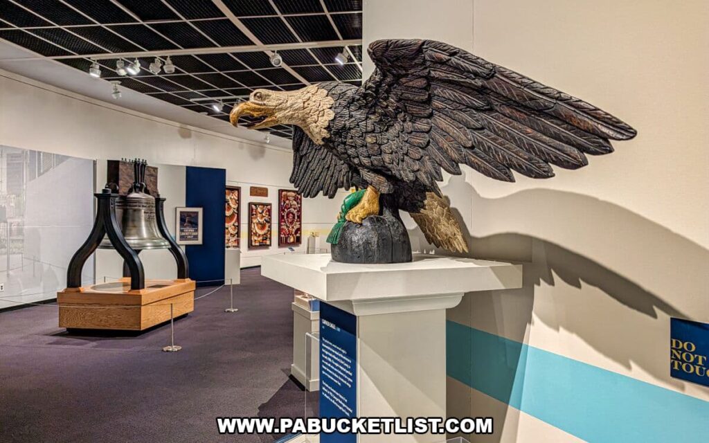 Large carved bald eagle sculpture displayed inside The State Museum of Pennsylvania in Harrisburg, shown with wings spread on a pedestal, with a Liberty Bell replica and Revolutionary War–themed exhibits visible in the background highlighting national symbols and Pennsylvania’s role in American history