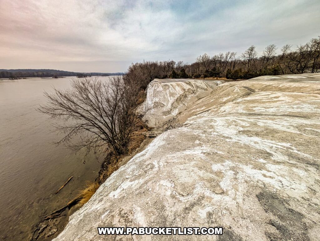 Chalky white quarry cliffs dropping toward the Susquehanna River at the White Cliffs of Conoy, with bare trees lining the riverbank under an overcast sky in Lancaster County, Pennsylvania.