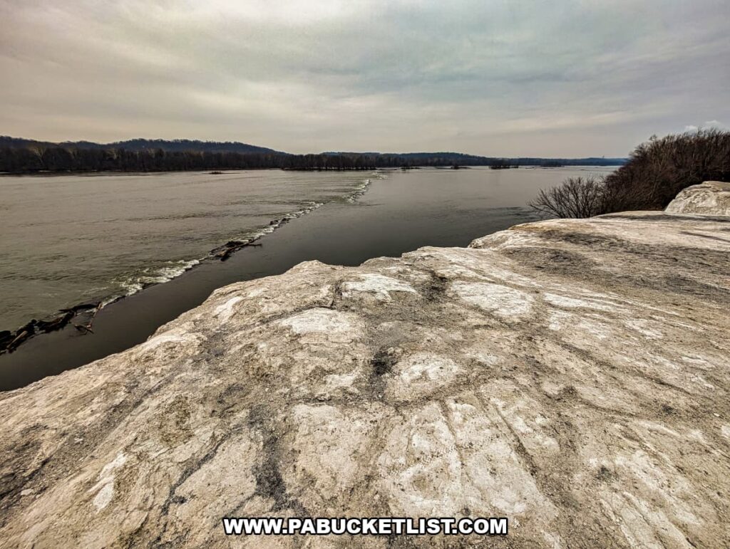 Chalky quarry surface at the edge of the White Cliffs of Conoy overlooking the Susquehanna River flowing westward, with distant wooded hills under an overcast sky in Lancaster County, Pennsylvania.