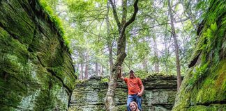 Two visitors stand among towering moss-covered rock formations at Bilger’s Rocks in Clearfield County, Pennsylvania, with massive tree roots winding over sandstone boulders in a lush forest setting.