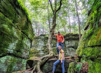 Two visitors stand among towering moss-covered rock formations at Bilger’s Rocks in Clearfield County, Pennsylvania, with massive tree roots winding over sandstone boulders in a lush forest setting.