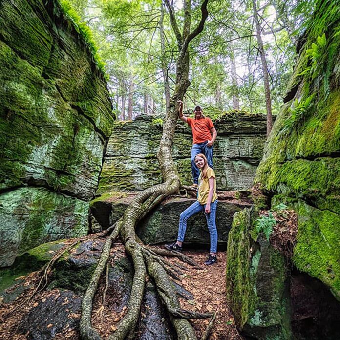 Two visitors stand among towering moss-covered rock formations at Bilger’s Rocks in Clearfield County, Pennsylvania, with massive tree roots winding over sandstone boulders in a lush forest setting.