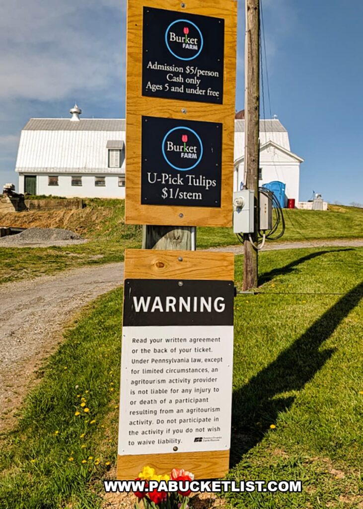 Wooden sign at Burket Farm in Blair County, Pennsylvania displaying admission details and U-pick tulip pricing, with a barn and farm buildings in the background