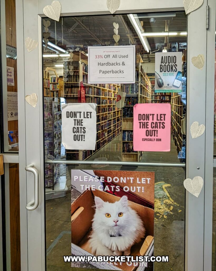 Front door of Cupboard Maker Books in Cumberland County, Pennsylvania, covered with signs reminding visitors not to let the cats out, including a poster of a fluffy white bookstore cat, with long rows of bookshelves visible inside the cozy Cat Bookstore.