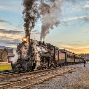 Steam locomotive No. 16 of the East Broad Top Railroad in Huntingdon County, Pennsylvania, billows smoke and steam as it pulls vintage passenger cars along the tracks at sunset.