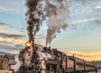 Steam locomotive No. 16 of the East Broad Top Railroad in Huntingdon County, Pennsylvania, billows smoke and steam as it pulls vintage passenger cars along the tracks at sunset.