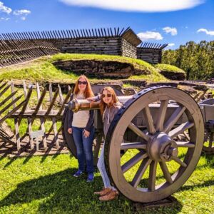 Two visitors pose beside a historic cannon at Fort Ligonier in Westmoreland County, Pennsylvania, with reconstructed wooden fort walls and grassy earthworks under a bright blue sky.