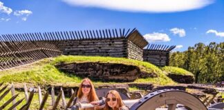 Two visitors pose beside a historic cannon at Fort Ligonier in Westmoreland County, Pennsylvania, with reconstructed wooden fort walls and grassy earthworks under a bright blue sky.