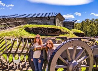 Two visitors pose beside a historic cannon at Fort Ligonier in Westmoreland County, Pennsylvania, with reconstructed wooden fort walls and grassy earthworks under a bright blue sky.