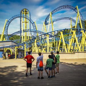 Group of kids watch the Impulse roller coaster at Knoebels Amusement Park in Columbia County, Pennsylvania, as the blue and yellow track twists and loops overhead on a sunny day.
