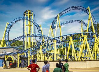 Group of kids watch the Impulse roller coaster at Knoebels Amusement Park in Columbia County, Pennsylvania, as the blue and yellow track twists and loops overhead on a sunny day.