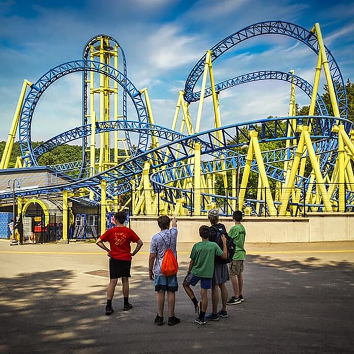 Group of kids watch the Impulse roller coaster at Knoebels Amusement Park in Columbia County, Pennsylvania, as the blue and yellow track twists and loops overhead on a sunny day.