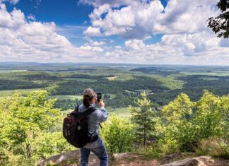 Hiker with a backpack stands at Indian Lookout in Huntingdon County, Pennsylvania, photographing sweeping views of forested ridges and farmland under a bright blue sky filled with puffy clouds.