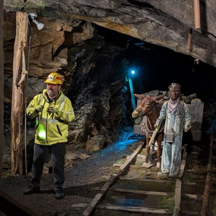 Lackawanna-Coal-Mine-Tour-Scranton-PA-square-crop (1) Tour guide in a hard hat leads visitors through the Lackawanna Coal Mine Tour in Scranton, Pennsylvania, passing historic mine rails and a display featuring a mule and miner statue deep inside the underground tunnel.