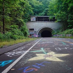 Entrance to the abandoned Rays Hill Tunnel along the Pennsylvania Turnpike in Bedford County, Pennsylvania, with colorful graffiti and chalk drawings covering the roadway leading into the dark tunnel beneath a wooded hillside.