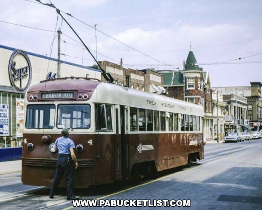 Color photograph of a vintage Philadelphia streetcar stopped on a city street, showing a transit worker standing at the front of the trolley, overhead electric wires, and storefronts and historic buildings lining the route in the background.