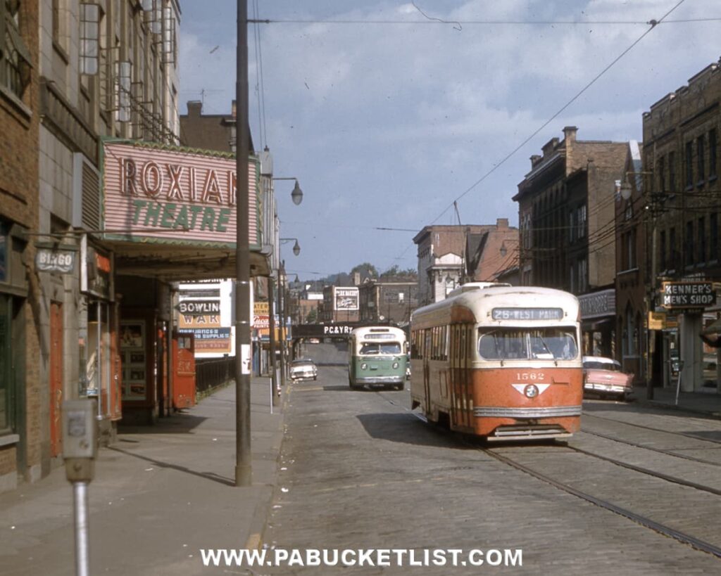 Historic street scene showing a vintage electric trolley traveling down a downtown Pennsylvania street past the Roxian Theatre marquee, with brick storefronts, overhead trolley wires, and additional streetcars and automobiles visible in the background.