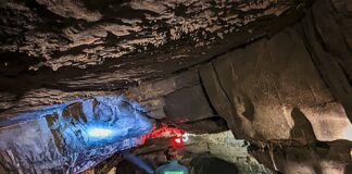 A visitor walks through a wide limestone passageway inside Woodward Cave in Centre County, Pennsylvania, illuminated by colorful cave lighting highlighting the low rock ceiling and textured walls.