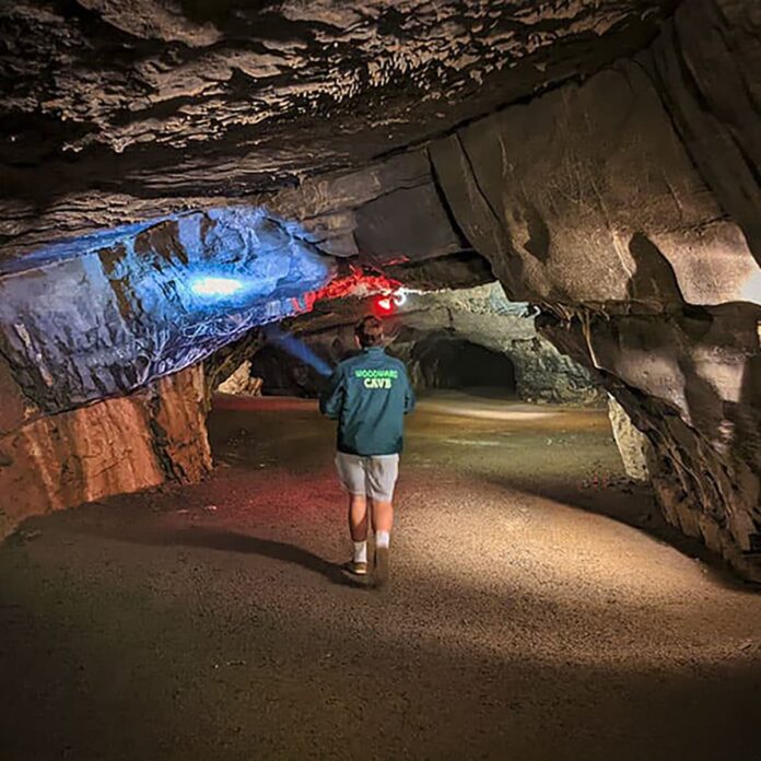 A visitor walks through a wide limestone passageway inside Woodward Cave in Centre County, Pennsylvania, illuminated by colorful cave lighting highlighting the low rock ceiling and textured walls.