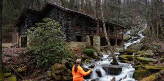 Hiker standing beside cascading water along Rattling Run at Acrigg’s Falls with Bordner Cabin overlooking the stream in Swatara State Park, Lebanon County, Pennsylvania.