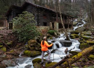 Hiker standing beside cascading water along Rattling Run at Acrigg’s Falls with Bordner Cabin overlooking the stream in Swatara State Park, Lebanon County, Pennsylvania.