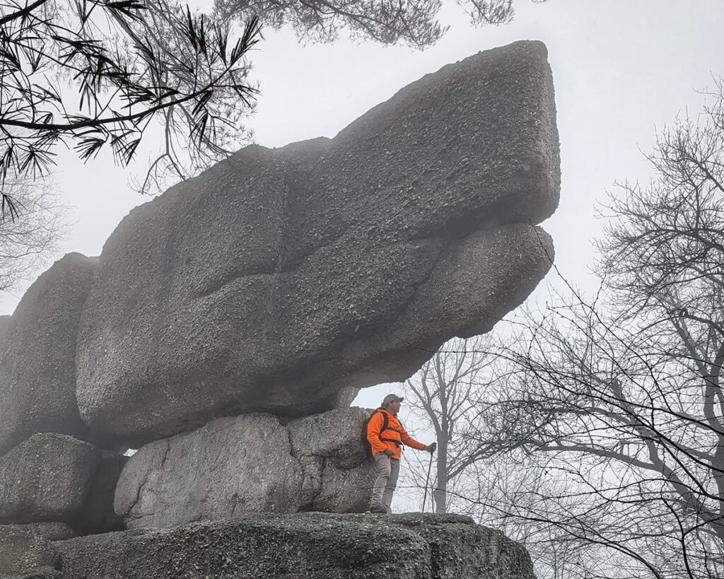 Hiker standing beneath a massive stacked conglomerate boulder at Boxcar Rocks on State Game Lands 211 in Lebanon County, Pennsylvania, with fog drifting through the surrounding forest.