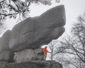 Hiker standing beneath a massive stacked conglomerate boulder at Boxcar Rocks on State Game Lands 211 in Lebanon County, Pennsylvania, with fog drifting through the surrounding forest.