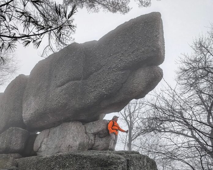Hiker standing beneath a massive stacked conglomerate boulder at Boxcar Rocks on State Game Lands 211 in Lebanon County, Pennsylvania, with fog drifting through the surrounding forest.