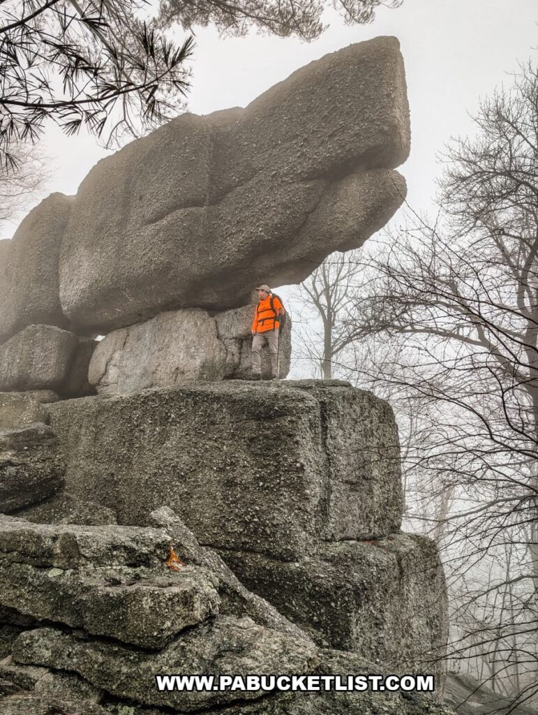 Hiker standing beneath massive stacked conglomerate boulders at Boxcar Rocks on Sharp Ridge in Lebanon County, Pennsylvania, with fog drifting through the surrounding forest.